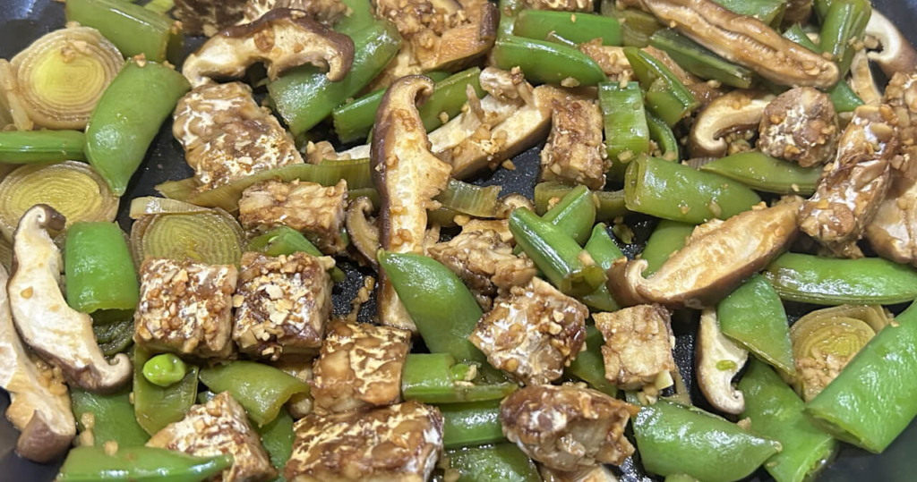 A photo of a stir fry made with no oil that includes tempeh, shiitake, leeks, and spring peas