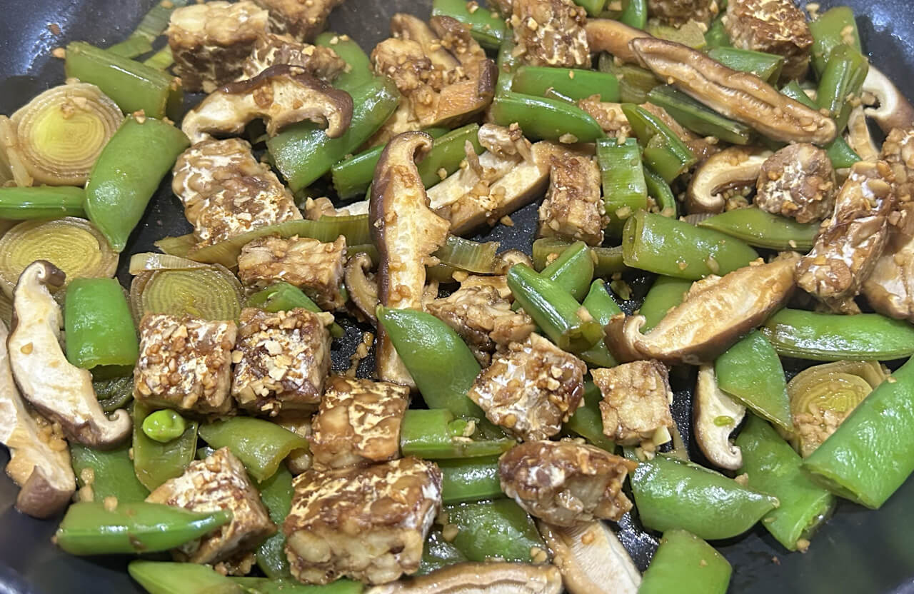 A photo of a stir fry made with no oil that includes tempeh, shiitake, leeks, and spring peas